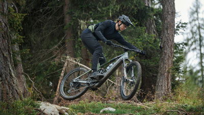 The image shows a man on a gray Cube Stereo Hybrid e-bike jumping over a slope in the forest.