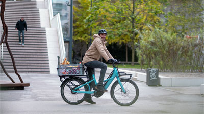 The image shows a man riding a blue Cube Longtail hybrid cargo bike through the city center with groceries stored on it.