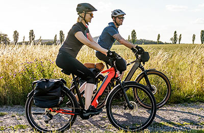 A couple riding e-bikes on a gravel path can be seen. Tall grass is visible in the background.