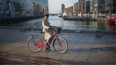 A woman is seen riding a pink Cube Ella Hybrid e-bike. She is riding along a river. A city can be seen in the background.