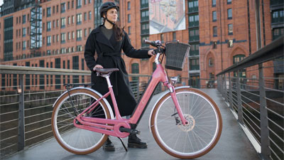 A woman is seen standing behind a pink Cube Ella Hybrid e-bike. A building is visible in the background.