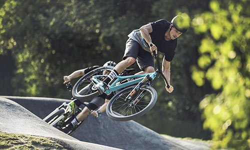 Cyclist jumps with a light blue dirt bike in a bike park, another cyclist with a green bike in the background.