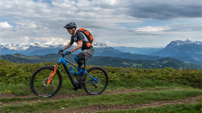 The image shows a man riding a blue Cube Stereo Hybrid e-bike with orange details. He is riding on a hiking trail with mountains visible in the background.