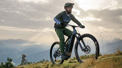 The image shows a man riding a gray Cube Stereo Hybrid e-bike on a hiking trail.
