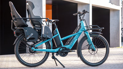 The photo shows a blue Cube Longtail hybrid cargo bike with two equipped child seats.