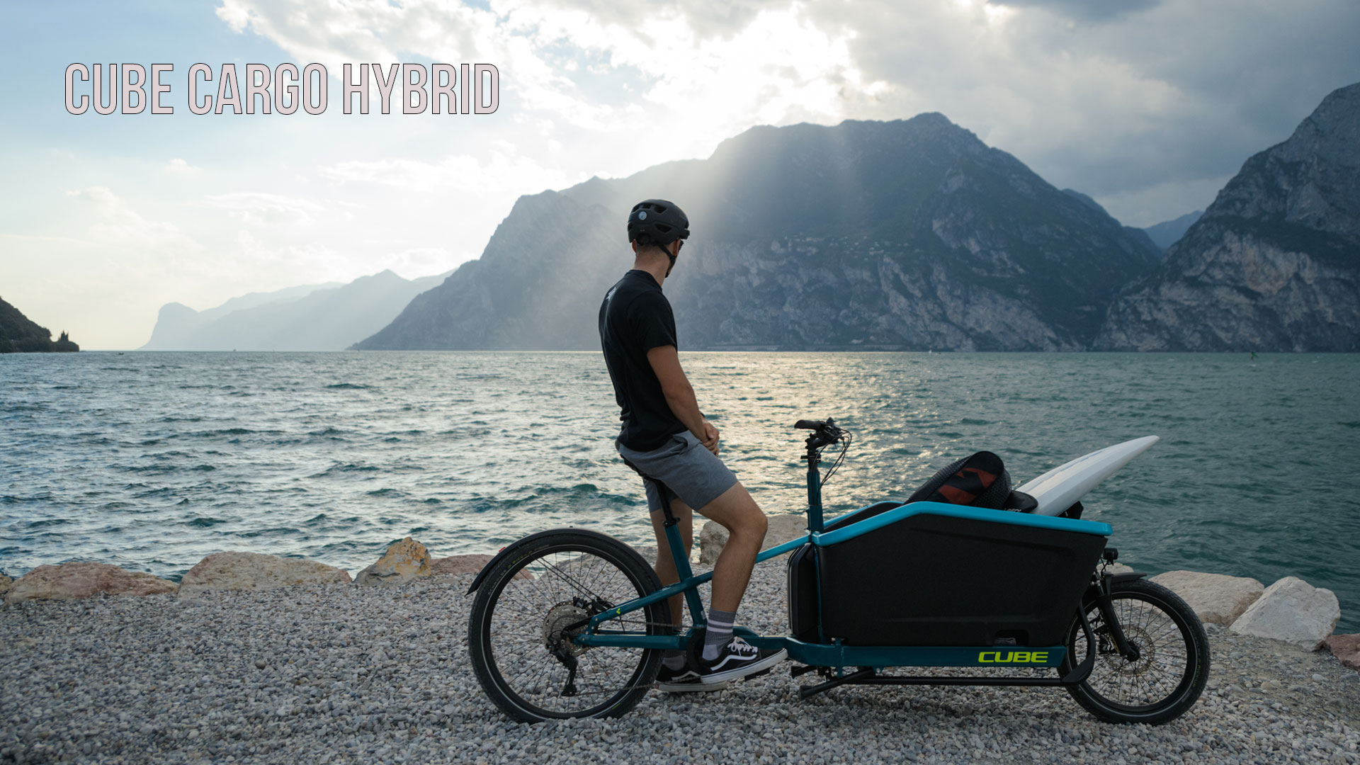 The image shows a man on a blue cargo bike standing on the shore of a lake, looking at the mountains in the background. The image is titled Cube Cargo Hybrid.