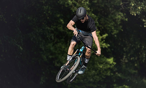 Cyclist jumps with a light blue dirt bike, trees can be seen in the background.