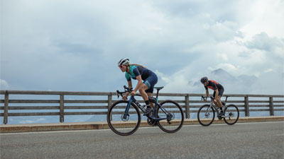 Two racing cyclists can be seen, with a wooden fence and clouds in the background.