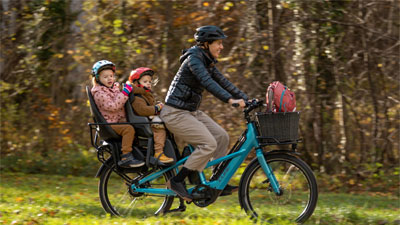 The image shows a mother on a blue Cube Longtail cargo bike. Two small children are sitting on the luggage rack.